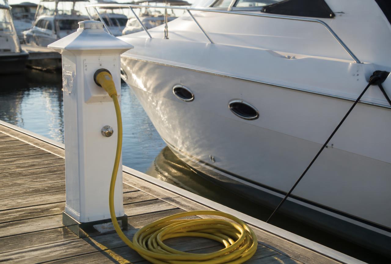 Yellow shore power cordset plugged into a marina dock pedestal beside a white motor yacht